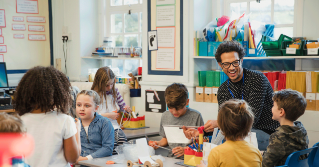 A male teacher in a classroom with young students, happily sharing the school supplies he bought from a company that was marketing to teachers with a teacher discount.