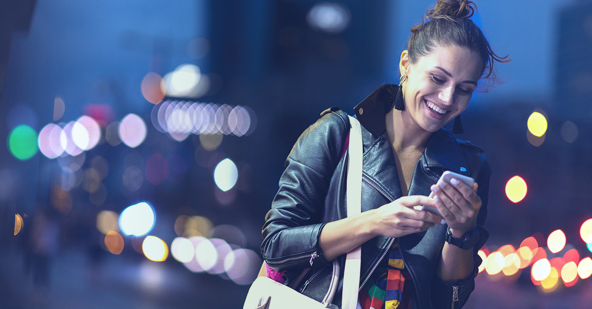 A woman on a street at night, smiling at her cell phone after receiving a personalized offer through an affiliate marketing program.
