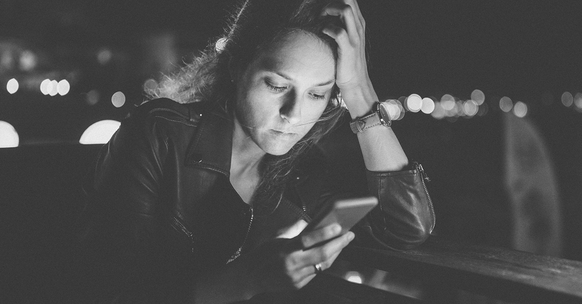 A concerned woman rests her head on her hands while she looks at her phone.