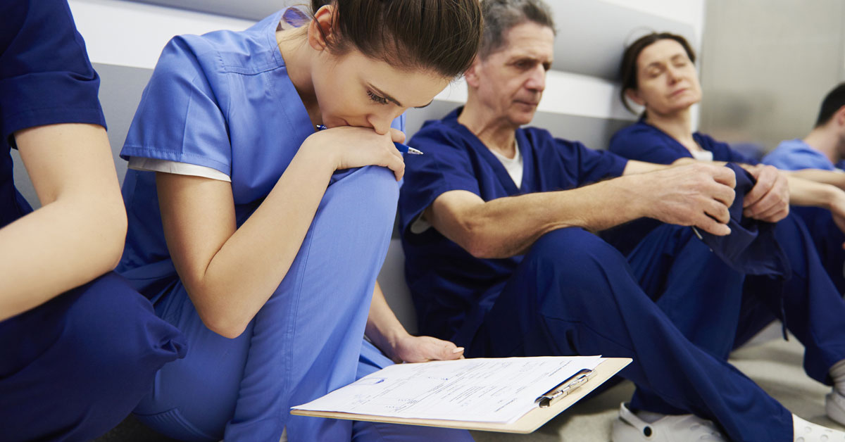 A worried nursing looking a clip while she sits in a hallway with other nurses and doctors.